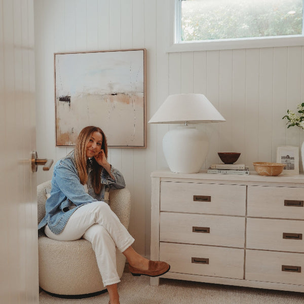 Woman sitting on a chair in a modern living room with a lamp and dresser.