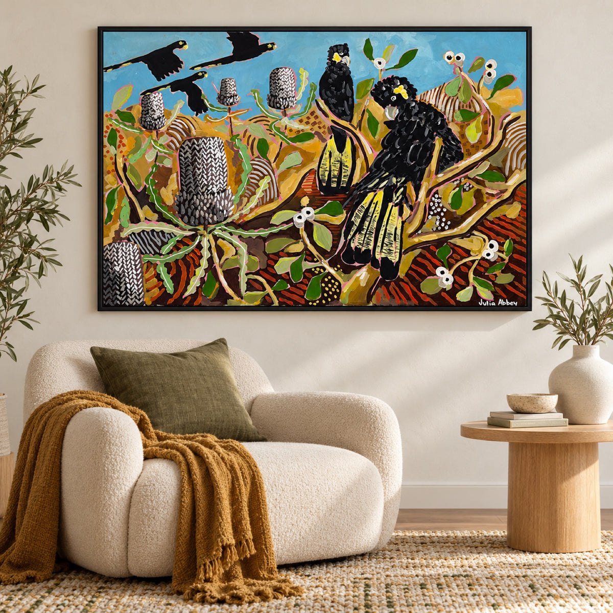 Basking In Banksias by Julia Abbey in a slim black frame above a cream lounge chair, styled with an olive cushion, ochre throw and oak side table.